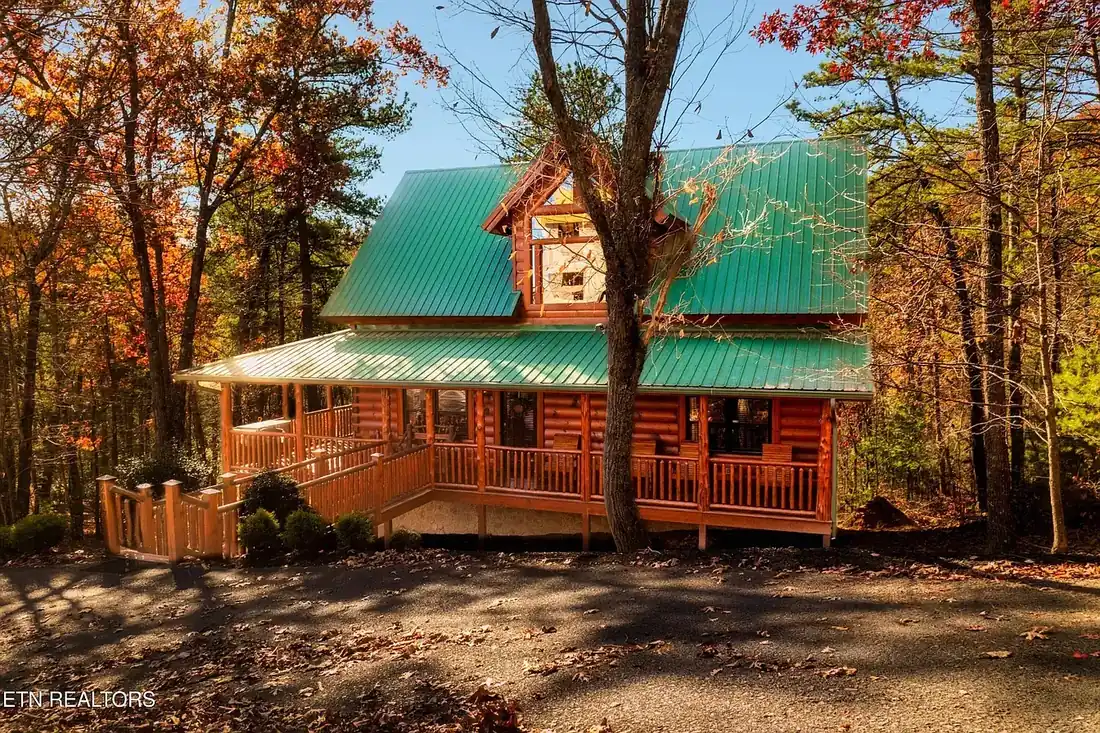 Front elevation of the two-storey log cabin with green metal roof, wraparound covered porch and log railings, surrounded by autumn trees