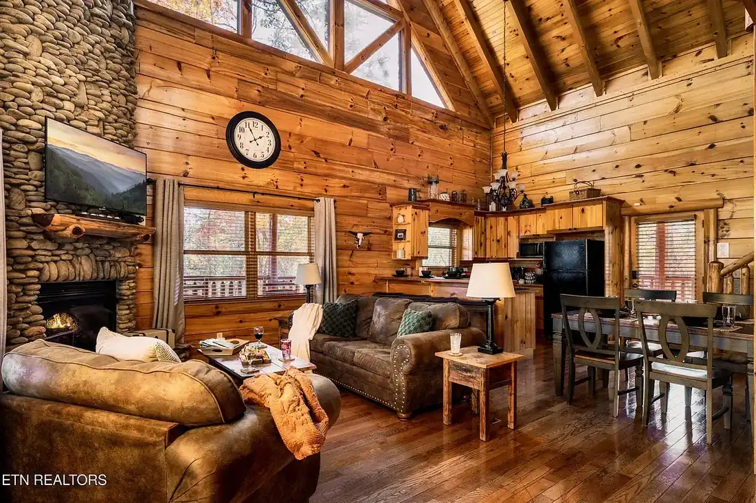 Great room with vaulted wood-beamed ceiling, prow windows, river rock fireplace, two sofas and the open kitchen and dining area beyond