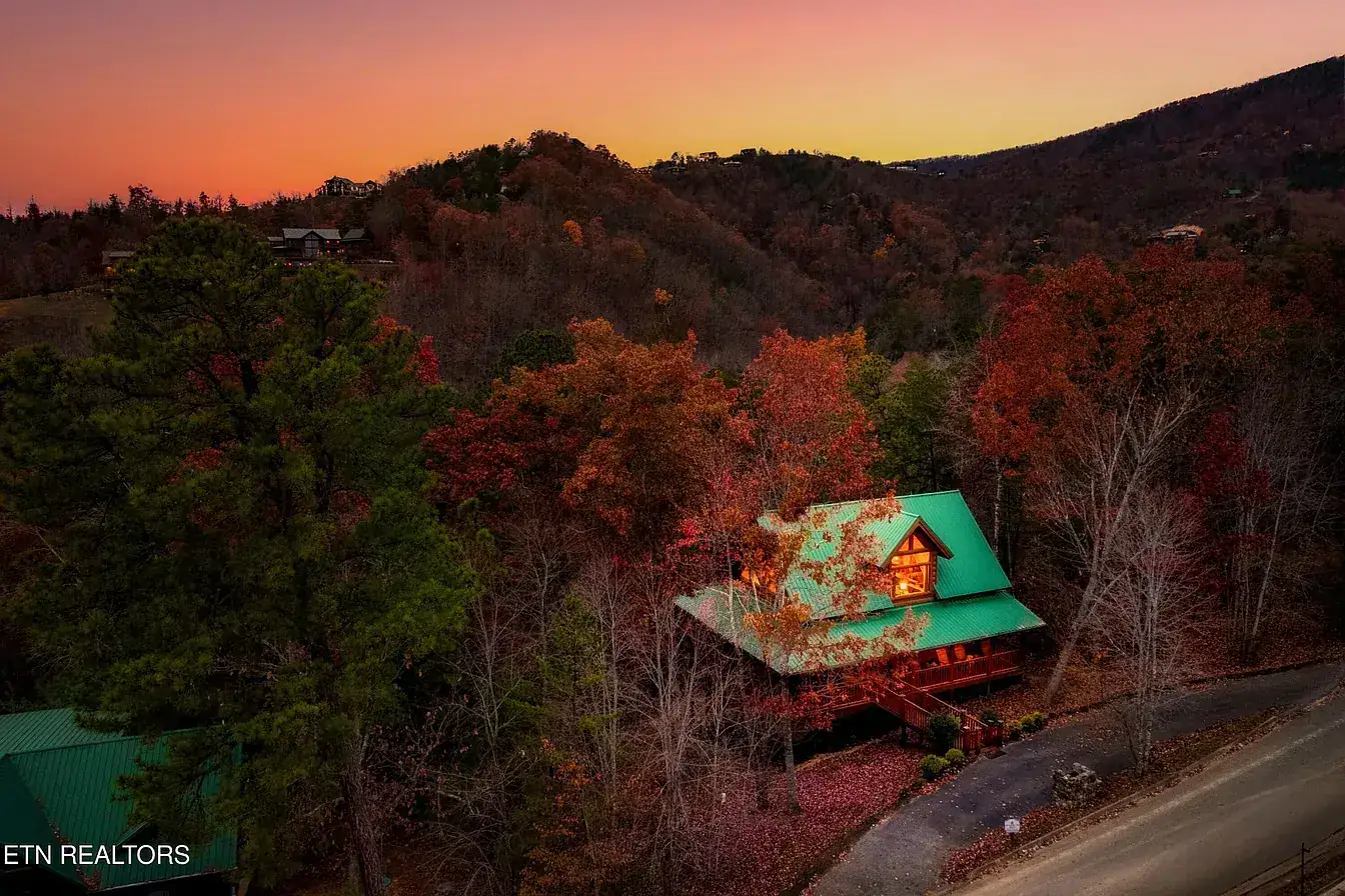 Aerial view at sunset of the log cabin with its green metal roof, tucked into a wooded hillside with autumn colour and the Smoky Mountain ridgeline behind